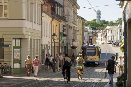 Belebte Steinstraße mit Fußgängern, Radfahrern, einer Straßenbahn und im Hintergrund die Friedenswarte auf dem Marienberg