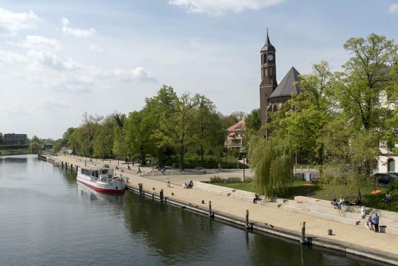 Salzhof-Ufer mit der Havel und einem Fahrgastschiff im Vordergrund, der St. Johanniskirche umgeben von Bäumen im Hintergrund